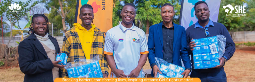 Catalyst Foundation Staff holding sanitary products with S.H.E Programme banners in the background. The banners state "SECURE HOPE EMPOWERED" and "Breaking barriers, transforming lives, inspiring dreams: Girls' rights matter!"
