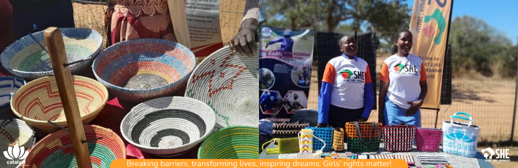 Two young women standing behind a table displaying a variety of woven baskets, bags, and other crafts at an outdoor event. The women are smiling and wearing t-shirts with "SHE" and "Catalyst Foundation" logos.