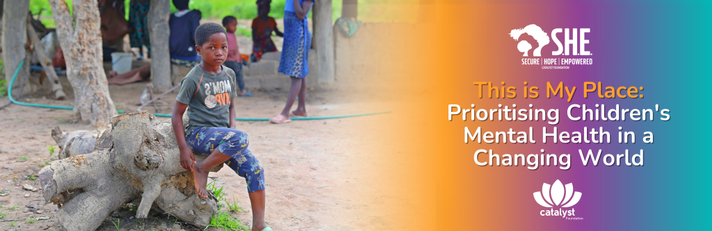 A wide banner for Children's Mental Health Week featuring a young boy sitting thoughtfully on a large fallen tree trunk in a rural village setting. To the right, a vibrant orange and purple gradient contains the text: "This is My Place: Prioritizing Children's Mental Health in a Changing World." The S.H.E. and Catalyst Foundation logos are prominently displayed at the top and bottom of the text section.